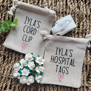 May include: Two small burlap drawstring bags with pink hearts and black text. The bags are labeled "IYLA'S CORD CLIP" and "IYLA'S HOSPITAL TAGS". A white plastic cord clip is also pictured.