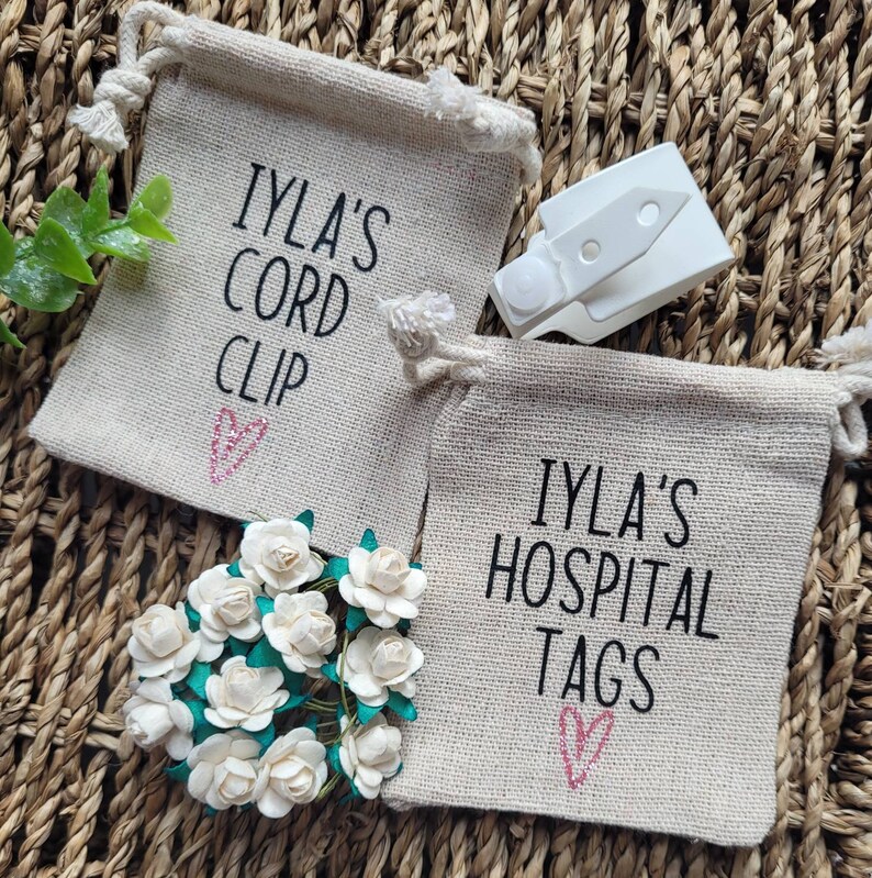 May include: Two small burlap drawstring bags with pink hearts and black text. The bags are labeled "IYLA'S CORD CLIP" and "IYLA'S HOSPITAL TAGS". A white plastic cord clip is also pictured.