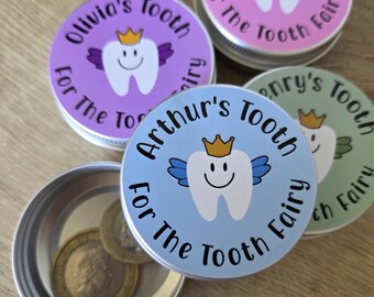 Könnte beinhalten: Vier runde Metalldosen mit Deckeln, die einen lächelnden Zahn mit goldener Krone und Flügeln zeigen. Jeder Deckel ist mit einem Namen und dem Text "For The Tooth Fairy" bedruckt. Eine geöffnete Dose enthält zwei goldfarbene Münzen.