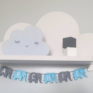 K&ouml;nnte beinhalten: Ein wei&szlig;es, wolkenf&ouml;rmiges Nachtlicht mit einem l&auml;chelnden Gesicht steht auf einem wei&szlig;en Regal mit einem grauen W&uuml;rfel. Unter dem Regal h&auml;ngt ein blaues und graues Elefantenbanner mit dem Namen "Archie" darauf.