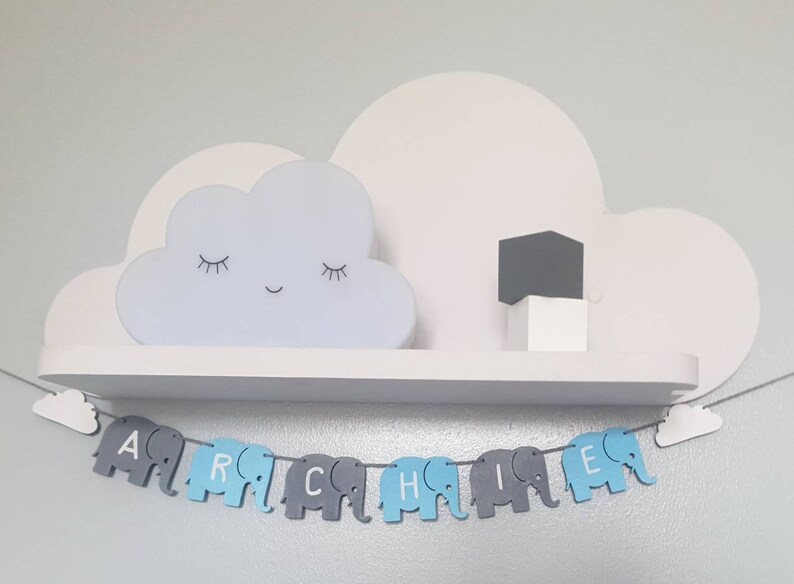 K&ouml;nnte beinhalten: Ein wei&szlig;es, wolkenf&ouml;rmiges Nachtlicht mit einem l&auml;chelnden Gesicht steht auf einem wei&szlig;en Regal mit einem grauen W&uuml;rfel. Unter dem Regal h&auml;ngt ein blaues und graues Elefantenbanner mit dem Namen "Archie" darauf.