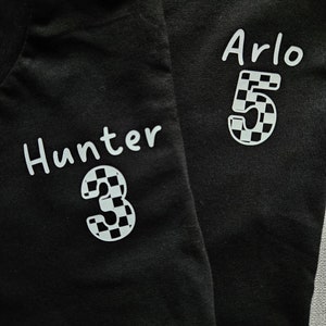 May include: Two black t-shirts with white text and numbers. The left shirt says "Hunter 3" and the right shirt says "Arlo 5". Both numbers are in a checkered pattern.