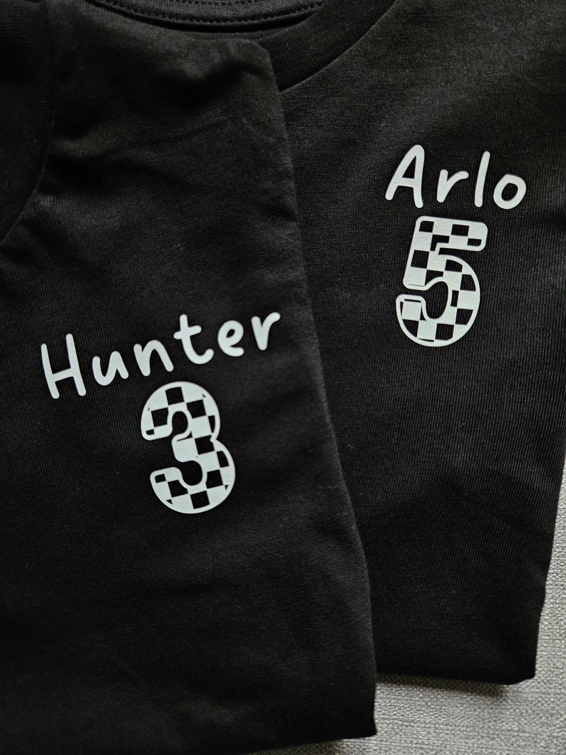 May include: Two black t-shirts with white text and numbers. The left shirt says "Hunter 3" and the right shirt says "Arlo 5". Both numbers are in a checkered pattern.
