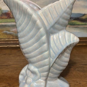 Può includere: Un vaso in ceramica azzurra a forma di foglia stilizzata. Il vaso ha una finitura lucida con sottili riflessi blu e rosa. Il design della foglia presenta venature dettagliate e un'apertura svasata. Altezza: circa 20 cm.