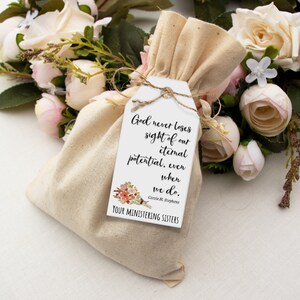 K&ouml;nnte beinhalten: Ein wei&szlig;es Etikett mit einem Blumenmuster und dem Text "God never loses sight of our eternal potential, even when we do. Carole M. Stephens YOUR MINISTERING SISTERS" befestigt an einem kleinen, beigefarbenen Stoffbeutel.