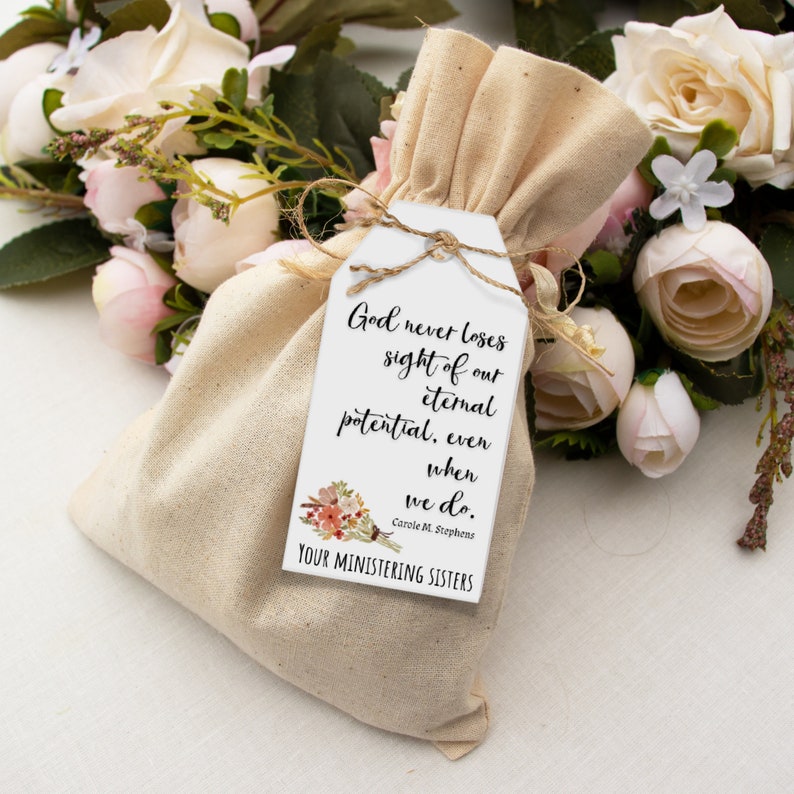 K&ouml;nnte beinhalten: Ein wei&szlig;es Etikett mit einem Blumenmuster und dem Text "God never loses sight of our eternal potential, even when we do. Carole M. Stephens YOUR MINISTERING SISTERS" befestigt an einem kleinen, beigefarbenen Stoffbeutel.