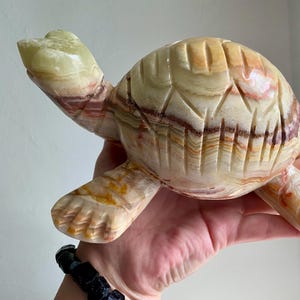Puede incluir: Una figura de tortuga de piedra tallada en tonos crema, beige y marrón. La tortuga tiene marcas detalladas en el caparazón y extremidades extendidas. La cabeza y las patas son de color verde claro. La figura se sostiene en una mano.