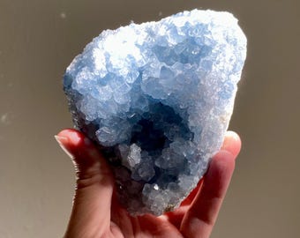 May include: A hand holds a large, light blue celestite crystal cluster. The crystal has a rough, textured surface with many small, crystalline formations. The colour is a soft, pale blue, with areas of deeper blue in the crevices.