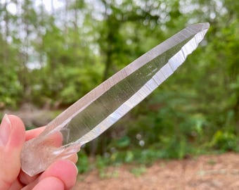 May include: A long, slender, clear quartz crystal point with a sharp, tapered tip. The crystal is transparent with visible internal striations and natural facets along its elongated body, showcasing a smooth, glassy texture throughout its length.