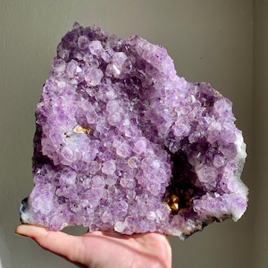 May include: A large, raw amethyst geode in shades of purple. The crystal structure is dense and sparkling, composed of many small, pointed crystals. The geode is held in a hand, highlighting its size and natural beauty.
