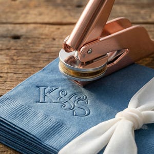 May include: A copper-colored embosser rests on a stack of blue napkins tied with a white ribbon. The napkins are embossed with the letters "K&S". The embosser is positioned over the napkins, ready to create an impression.