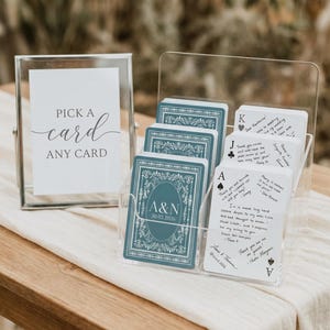 May include: A clear acrylic card holder with a stack of teal playing cards featuring a decorative design and the initials "A&N" with a date. A framed sign reads "Pick a card, any card."