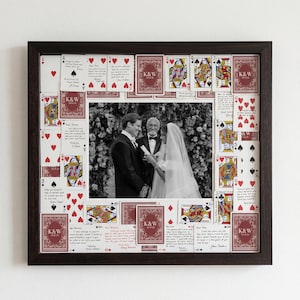May include: A framed wedding photo surrounded by playing cards. The frame is dark brown, and the photo is in black and white. The playing cards are red and white, with some featuring the initials "K&W".