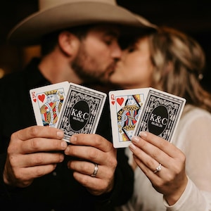May include: Playing cards featuring a King of Hearts and a Queen of Hearts, held by a couple. The cards have a personalized design on the back with the initials "K&C" and the date "06.06.26". The couple is kissing in the background.