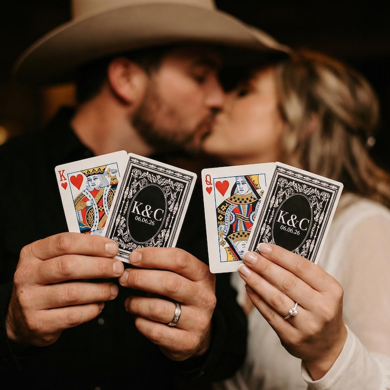 May include: Playing cards featuring a King of Hearts and a Queen of Hearts, held by a couple. The cards have a personalized design on the back with the initials "K&C" and the date "06.06.26". The couple is kissing in the background.