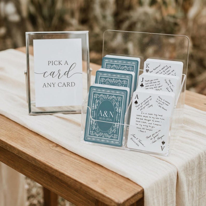 May include: A clear acrylic card holder with a deck of teal playing cards featuring a floral design and the initials "A&N" with a date. A sign in a silver frame reads "Pick a card, any card."