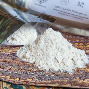 Peut inclure: Un tas de poudre claire est présenté sur une surface décorative. Un sachet argenté, partiellement ouvert, révèle plus de poudre. L'étiquette indique 100% Lambda Carraghénane, avec un poids net de 228 g.