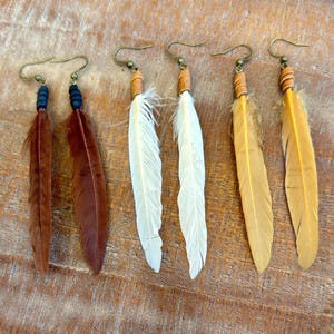 May include: Four pairs of feather earrings with leather and metal accents. The earrings are in brown, white, and yellow.