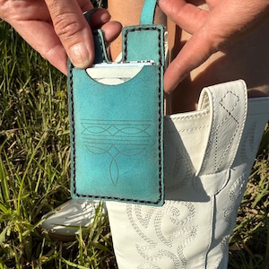 Könnte beinhalten: Ein türkisfarbener Lederkartenhalter mit einem dekorativen Stichdesign und schwarzen Nähten an den Kanten. Der Kartenhalter hat eine Schlaufe oben und wird in der Nähe eines weißen Cowboy-Stiefels gehalten.