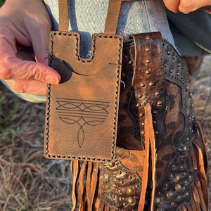 May include: A brown leather phone case with a decorative stitched design and a strap, held near a brown leather cowboy boot with fringe and metal studs. The case has a U-shaped top and a decorative stitched design.