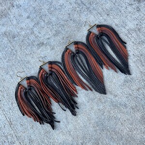 May include: Three pairs of leather fringe earrings in black, brown, and tan. The earrings are all different sizes, with the largest pair being the longest. The earrings are hanging from a gold-colored metal hook.