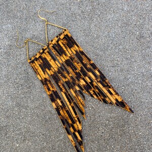 Op de afbeelding: Een paar lange, gouden, driehoekige oorbellen met een luipaardprint franje.