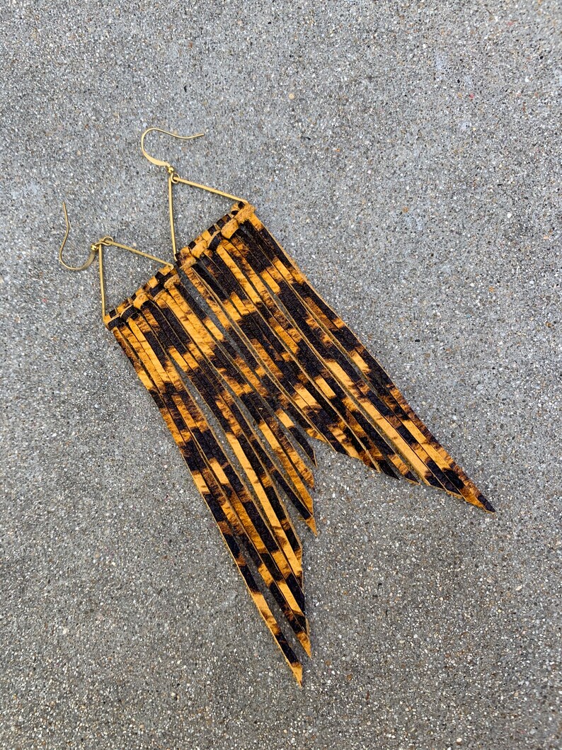 Op de afbeelding: Een paar lange, gouden, driehoekige oorbellen met een luipaardprint franje.