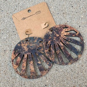 May include: A pair of large, round, wooden earrings with a sunburst design. The earrings are brown and black with a distressed finish. The earrings are hanging from gold-toned hooks.