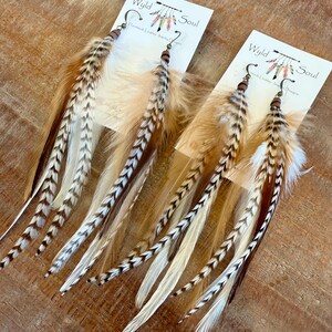 May include: A pair of long feather earrings with brown, white, and tan feathers. The earrings are handmade and have a wooden bead and a brass hook. The earrings are labeled "Wyld Soul Handmade Leather Jewelry"