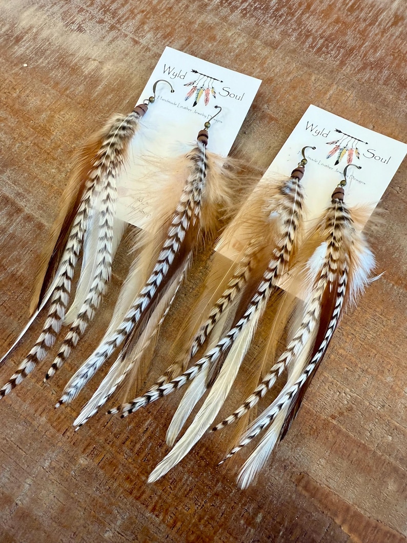 May include: A pair of long feather earrings with brown, white, and tan feathers. The earrings are handmade and have a wooden bead and a brass hook. The earrings are labeled "Wyld Soul Handmade Leather Jewelry"