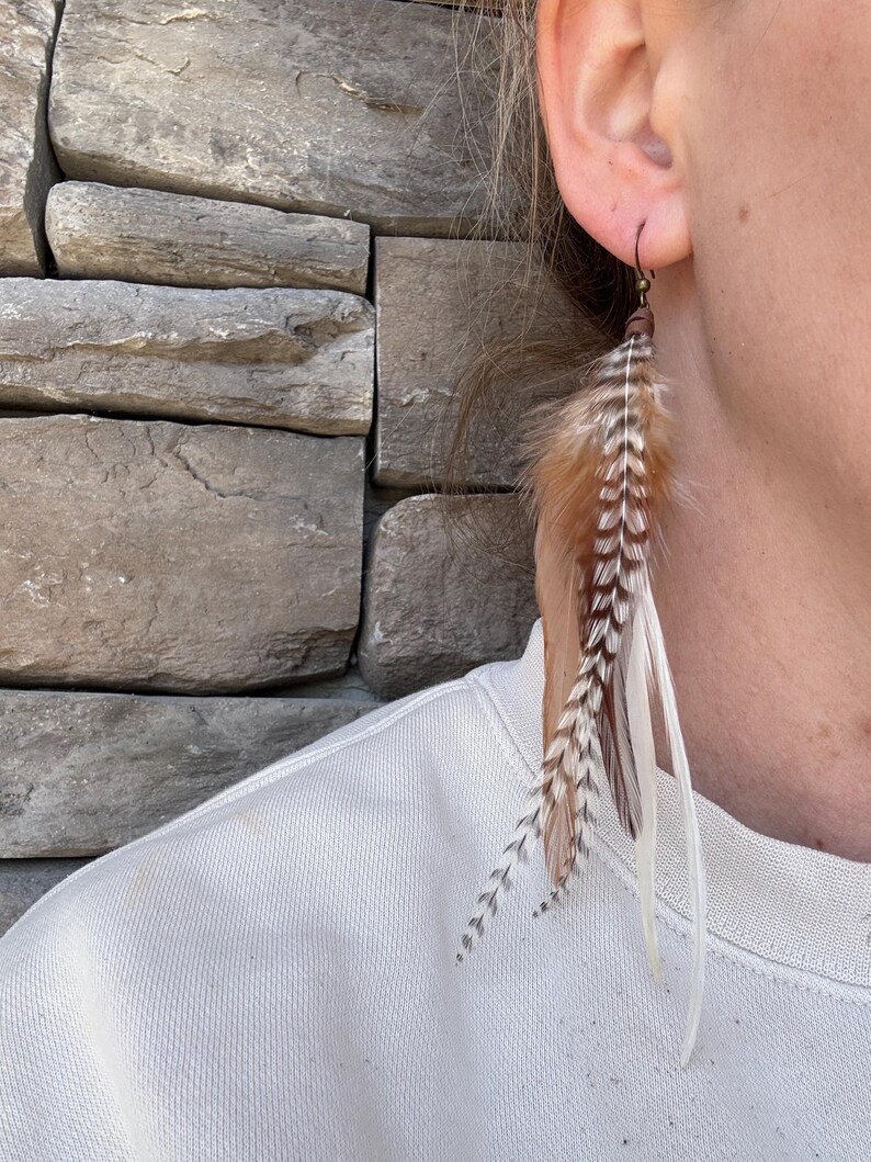 May include: A pair of long feather earrings with brown, white, and black feathers. The earrings are hanging from a person's ear.