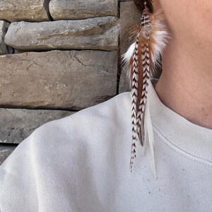 May include: A pair of long, brown and white feather earrings with a black hook. The earrings are hanging from a person's ear.