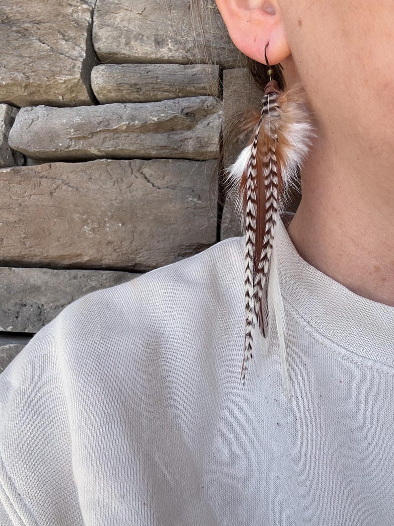 May include: A pair of long, brown and white feather earrings with a black hook. The earrings are hanging from a person's ear.