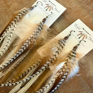 May include: A pair of long, brown and white feather earrings with brown leather and metal accents. The earrings are labeled "Wyld Soul" and "Handmade Leather Jewelry Designs".