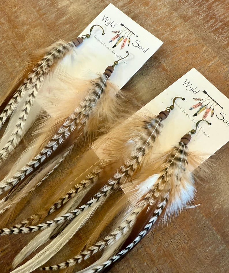 May include: A pair of long, brown and white feather earrings with brown leather and metal accents. The earrings are labeled "Wyld Soul" and "Handmade Leather Jewelry Designs".