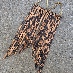 Op de afbeelding: Een paar gouden driehoekige oorhangers met lange bruine en zwarte luipaardprint franjes.
