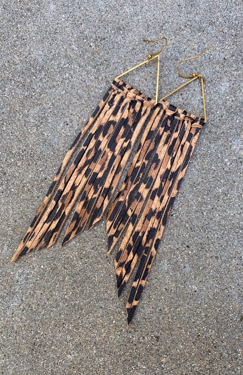 Op de afbeelding: Een paar gouden driehoekige oorhangers met lange bruine en zwarte luipaardprint franjes.