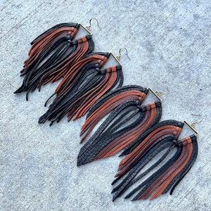 May include: Three pairs of leather fringe earrings in black, brown, and tan. The earrings are made with a square-shaped metal frame and have long, flowing fringe.