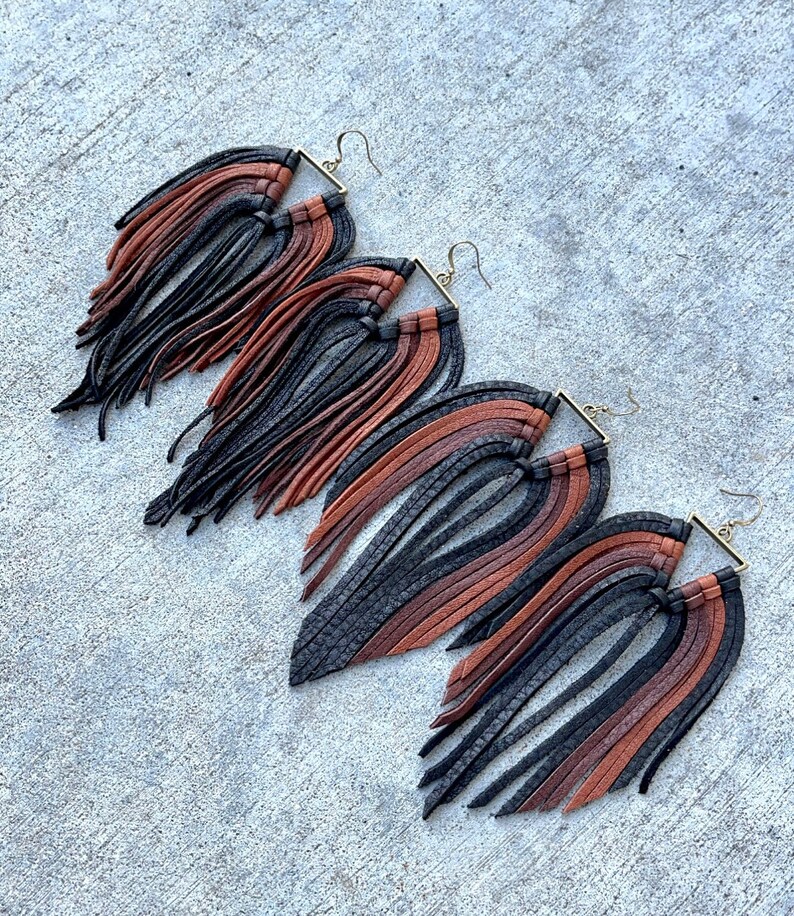 May include: Three pairs of leather fringe earrings in black, brown, and tan. The earrings are made with a square-shaped metal frame and have long, flowing fringe.