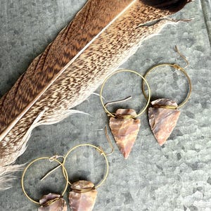 May include: A pair of gold hoop earrings with a pink arrowhead pendant. The arrowheads are wrapped in gold wire and have a natural, rough texture. The earrings are displayed on a gray background with a brown feather.