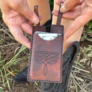 May include: A brown leather card holder with a decorative stitched design. The card holder has a vertical design with a strap on each side. The card holder is held in front of a pair of black cowboy boots.