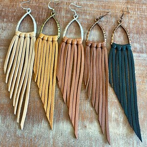 May include: Five pairs of leather fringe earrings with a teardrop-shaped metal top. The earrings are in various colors, including tan, brown, and black.