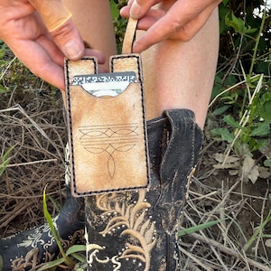 May include: A tan leather cardholder with a decorative stitched design and a small strap. The cardholder is held up, revealing cards and cash inside. The background includes a pair of black cowboy boots with gold floral embroidery.