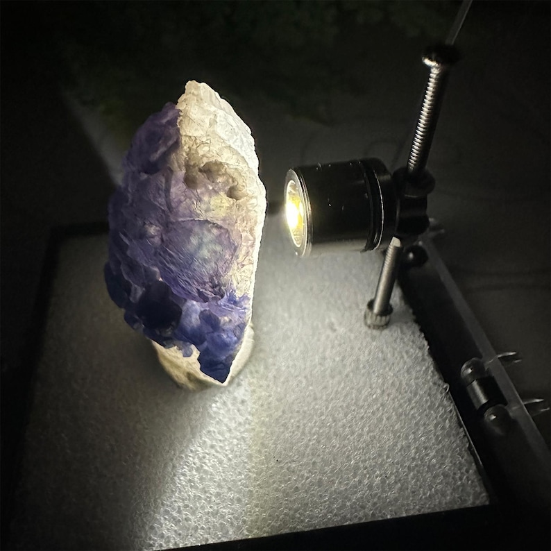 May include: A rough-hewn, natural stone specimen, predominantly white and deep purple, illuminated by a focused light source. The stone's surface has a textured appearance, with areas of color variation. The lighting highlights the stone's unique form and details.