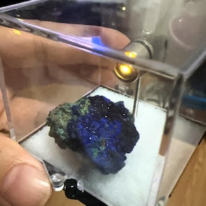 May include: A blue and green mineral specimen displayed in a clear acrylic case. The mineral has a crystalline structure with a deep blue color, accented by patches of green. The case is illuminated by a small light.