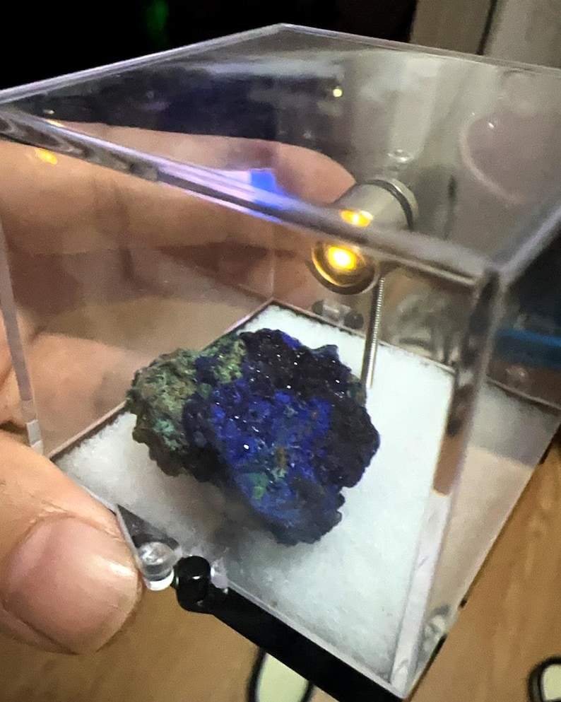 May include: A blue and green mineral specimen displayed in a clear acrylic case. The mineral has a crystalline structure with a deep blue color, accented by patches of green. The case is illuminated by a small light.