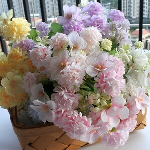 Könnte beinhalten: Ein Strauß künstlicher Blumen in einem geflochtenen Korb. Die Blumen sind in den Farben Rosa, Gelb und Lila.