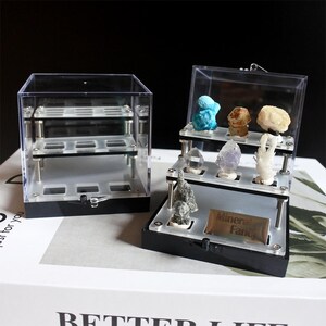 May include: Two clear acrylic display cases for mineral specimens. One case is a cube with three shelves. The other case is a tiered display with three shelves. The tiered display has a label that says "Mineral Fancy".