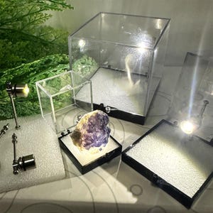 May include: A collection of clear acrylic display cases and small spotlights. A purple and white mineral specimen is illuminated inside a black-based display case. The scene is set against a backdrop of greenery and a patterned surface.