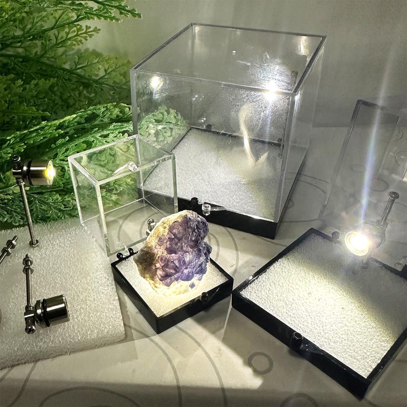 May include: A collection of clear acrylic display cases and small spotlights. A purple and white mineral specimen is illuminated inside a black-based display case. The scene is set against a backdrop of greenery and a patterned surface.